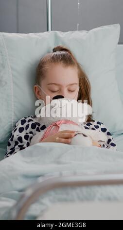 Portrait of tired sick child sleeping after suffering medical recovery surgery during disease examination in hospital ward. Hospitalized kid resting in bed wearing oxygen nasal tube Stock Photo