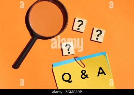 Top view of magnifying glass, wooden cube with question mark and color notepaper with text Q and A on orange background. Stock Photo