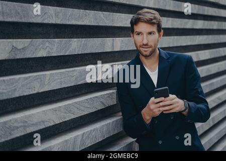 Thoughtful young bearded man standing outside holding mobile, typing on phone keyboard Stock Photo