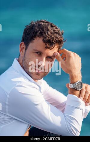 Side view of self assured handsome male wearing white shirt and wristwatch leaning on hand and looking at camera on blurred background of sea on sunny day Stock Photo