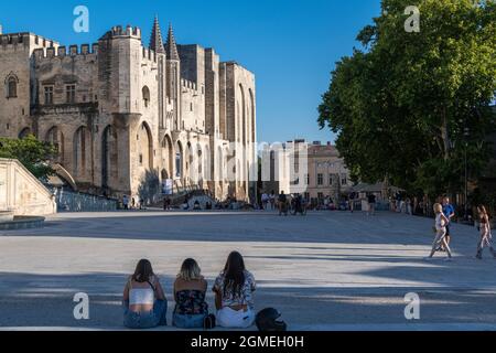 3 girls sit looking across Place du Palais towards the Palais des Papes, Avignon, France. Stock Photo