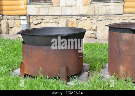 two large black metal cast-iron cauldrons for pilaf on a homemade stand from a large pipe outside on green grass Stock Photo
