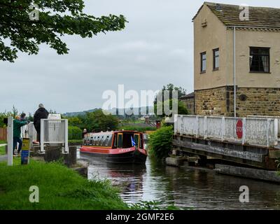 People watch hire narrowboat travelling past & through Micklethwaite swingbridge, barriers open - Leeds Liverpool Canal, Yorkshire, England, UK. Stock Photo