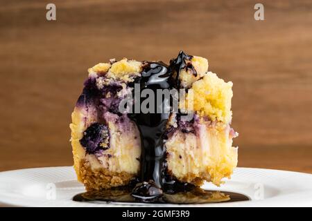 Closeup view slice of delicious homemade blueberry and crumble cheesecake poured with chocolate syrup in white ceramic plate on wooden table. Stock Photo