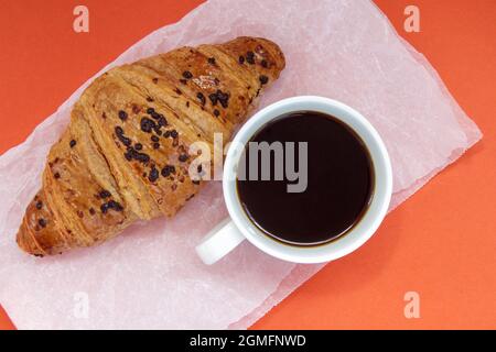 Black coffee without milk in a white cup and a chocolate croissant on parchment and bright background. French breakfast with fresh pastries. Top view Stock Photo