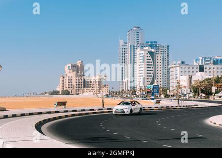 Toyota Taxi Car Moving In Street Of City Of Ajman. Building Of Bahi Ajman Palace Hotel In Sunny Day.Urban Architecture Of UAE Resort Town Of Ajman Stock Photo