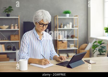 Mature woman sitting at her desk at home, using tablet and taking notes in notebook Stock Photo