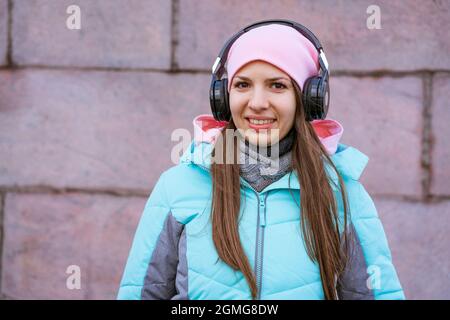 Happy young woman in warm jacket and pink hat listening to music on headphones against the background of a granite wall, travel among the city streets Stock Photo