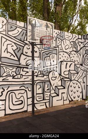 A basketball basket and backboard stand on a sports field against a painted wall with graffiti and trees. Stock Photo