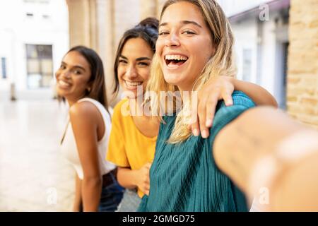 Portrait of three cheerful multiethnic female friends taking selfie with phone on city street - Friendship and millennial people concept Stock Photo