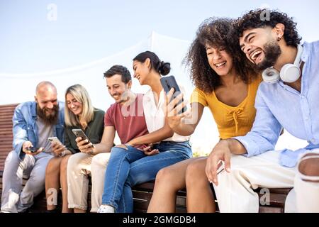 Diverse group of happy college friends using mobile phone while sitting together outdoors - Young students watching sharing content on social media Stock Photo