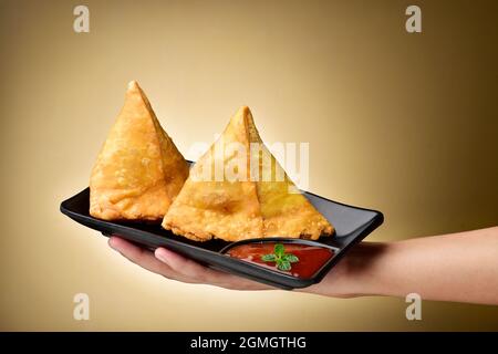 Samosa plate in hand, hand holding plate with  samosa and tomato ketchup over brown background Stock Photo
