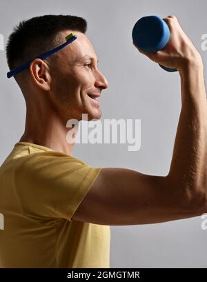 Portrait of smiling adult man in yellow t-shirt holding toothbrush behind his ear and a dumbbell in hand. Side view Stock Photo
