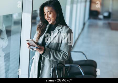 Woman waiting at airport lounge using her mobile phone. Smiling business traveler at airport waiting for the flight. Stock Photo