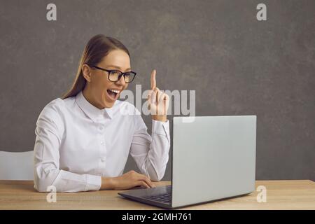 Young happy woman sitting at desk with raised hand finger working on laptop Stock Photo