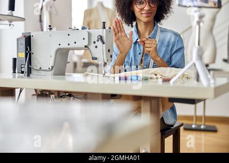 Smiling African-American seamstress shows black thread to camera sitting at workplace with sewing machine Stock Photo