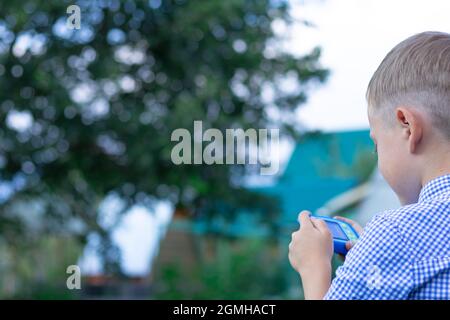 A cute preschool boy with a neat hairstyle in a blue shirt takes pictures of green plants on a hot summer day. Selective focus. Portrait Stock Photo