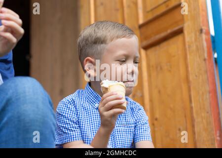 Contented cheerful child boy in a blue shirt eats ice cream on the porch of a house in a village on a summer sunny day. Selective focus. Portrait Stock Photo