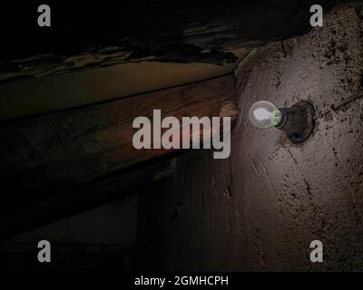 Light bulb glowing dimly in an abandoned old basement with wooden beams and stone wall Stock Photo
