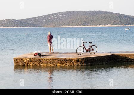 Vodice, Croatia - August 16, 2021: Young man standing alone on a sea pier and fishing with his bike parked next to him Stock Photo