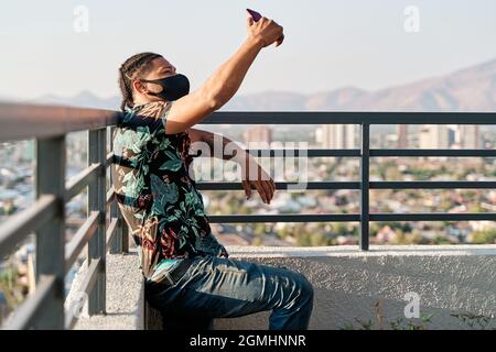 young latin man taking a selfie with a mask on the roof of the building, concept of social distancing and technology. Stock Photo