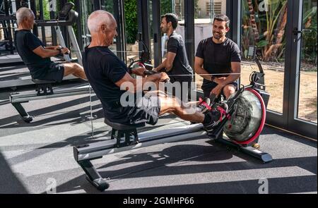 An elderly European man, in a gym, with a personal trainer, exercising simulating rowing, on a simulator, indoor. Stock Photo