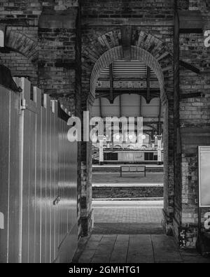 A railway station platform and a locomotive can be seen through an archway.  There is a white wooden partition on one side. Sunlight falls on the plat Stock Photo