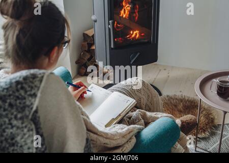 Young woman write in notebook sitting in armchair by fireplace Stock Photo