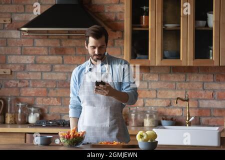 Bearded man using smartphone, distracted from cooking in kitchen Stock Photo