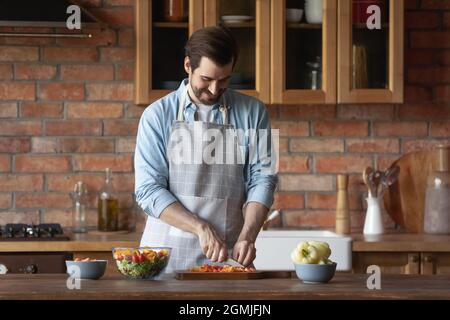 Smiling man in apron cutting vegetables, cooking at home Stock Photo