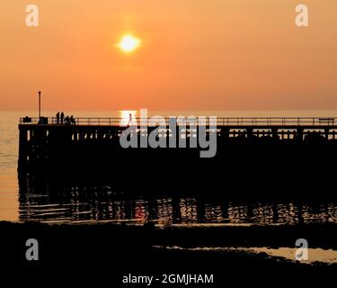 Hazy orange sunset on Welsh coast (Aberystwyth, Wales, UK) over a silhouetted pier with people. Person captured in sun's glow. Stock Photo