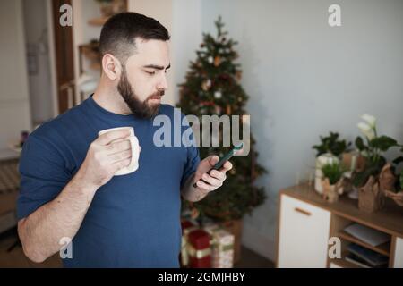 Serious young Caucasian man with beard standing with mug in living room with Christmas tree and checking message on phone Stock Photo