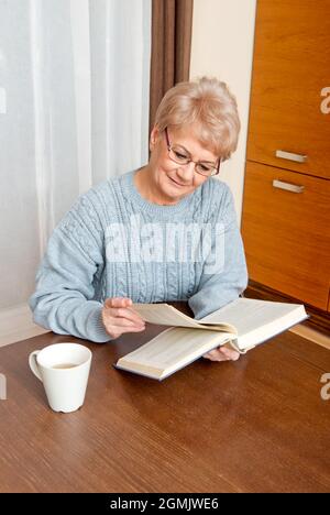 An elderly woman reading a book and drinking coffee at home Stock Photo