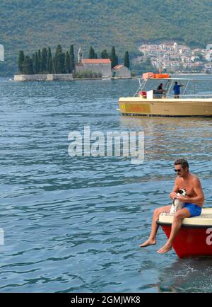 A man with his small dog sits at the bow of a small motor boat on the calm waters of Kotor Bay. Stock Photo