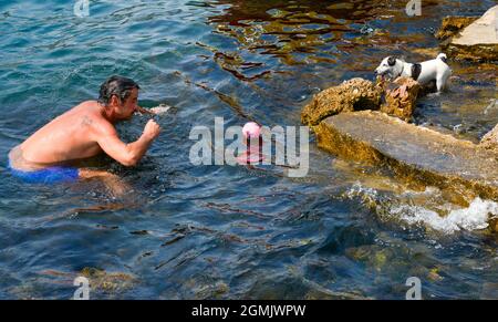 A man in swimming shorts plays with his small pet dog with a plastic ball,in the seawater on a summer afternoon.