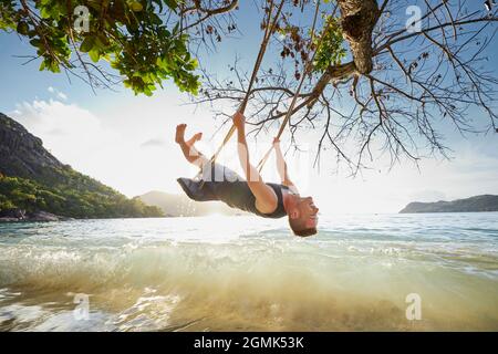 Happy man on swing enjoying holiday. Beautiful sunset on beach in Seychelles. ​ Stock Photo