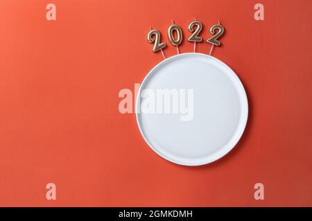 Creative Christmas table setting. Empty plate with straight edges, candles in form of numbers 2022, on red background, copy space, minimal layout, moc Stock Photo