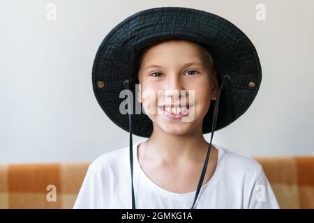 Candid shot of happy child with smiling face sitting on sofa, wearing white t-shirt and black panama hat. Portrait of a positive caucasian child sitting alone relaxing at home on weekend Stock Photo