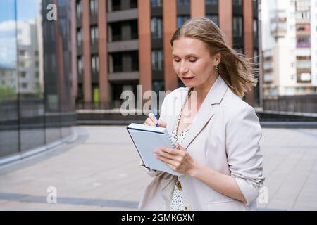 Business woman in light jacket stands and writes something near business center. Caucasian girl writes with pen in notebook goals plans affairs and list of wishes for work. Notes of successful lady Stock Photo