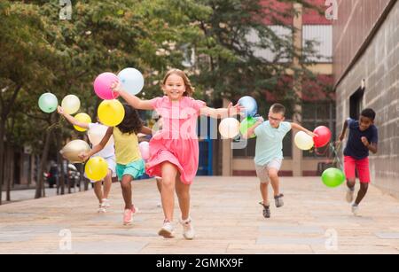 Happy tweens wth colorful balloons chasing each other on city street ...