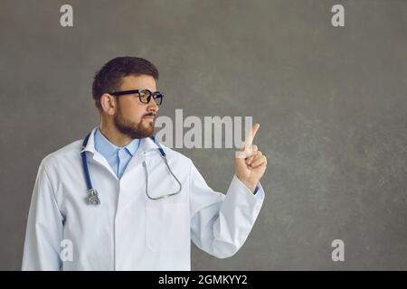 Handsome young doctor isolated on gray background points finger at copy space for text Stock Photo