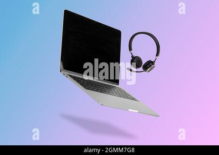 Laptop  computer with empty screen and headset  levitating in the air on a background illuminated by a blue and pink color gradient. Creative minimal Stock Photo