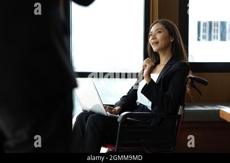 Attractive disabled businesswoman in wheelchair talking with her manager, working on laptop while meeting Stock Photo