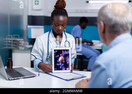 African american medic holding virus animation on tablet screen for old patient in healthcare office. Black doctor showing bacteria results from professional examination and consultation Stock Photo