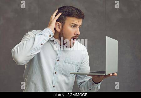 Shocked young man looking at screen of laptop computer that he's holding in hand Stock Photo