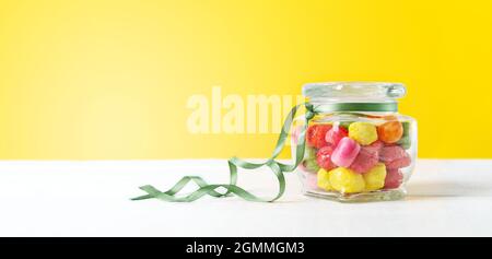 Glassware with different chewy multi-colored candies in the form of fruits, with a yellow backdrop. Banner Stock Photo