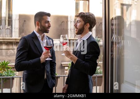Businessmen in suite talking together against a window in an hotel room holding red wine glasses. Stock Photo
