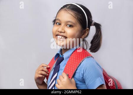 Portrait of a happy smiling Indian girl child student in blue school uniform with red bag on grey background Stock Photo