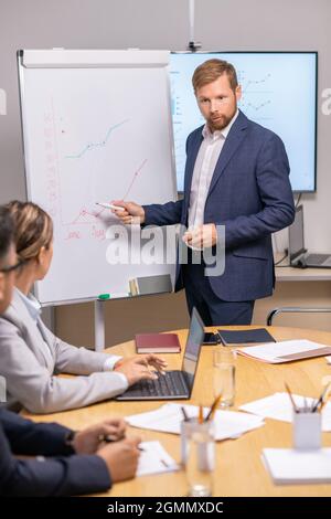 Young serious businessman pointing at graph on whiteboard and looking at economists during presentation Stock Photo