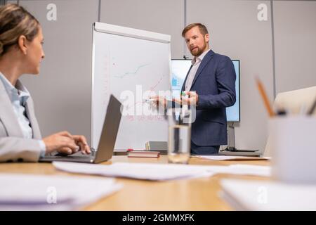 Contemporary coach pointing at whiteboard at seminar while looking at businesswoman with laptop Stock Photo
